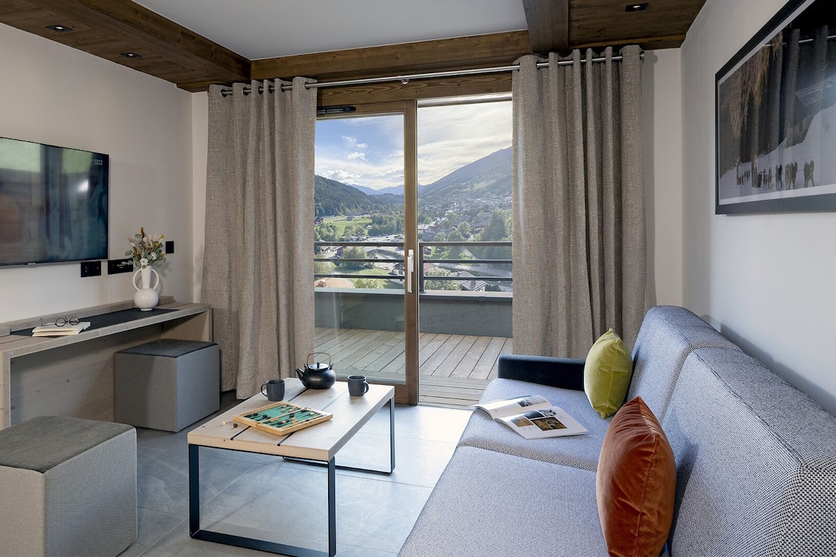 A modern living room with a flat-screen TV, a sofa, a coffee table, and a balcony with a mountain view.