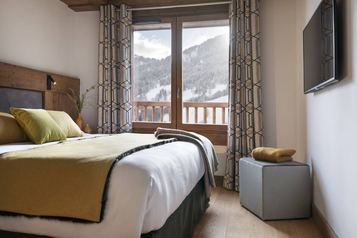 A bedroom with a bed, a small bedside table, a nightstand, a grey ottoman, and a window with curtains.