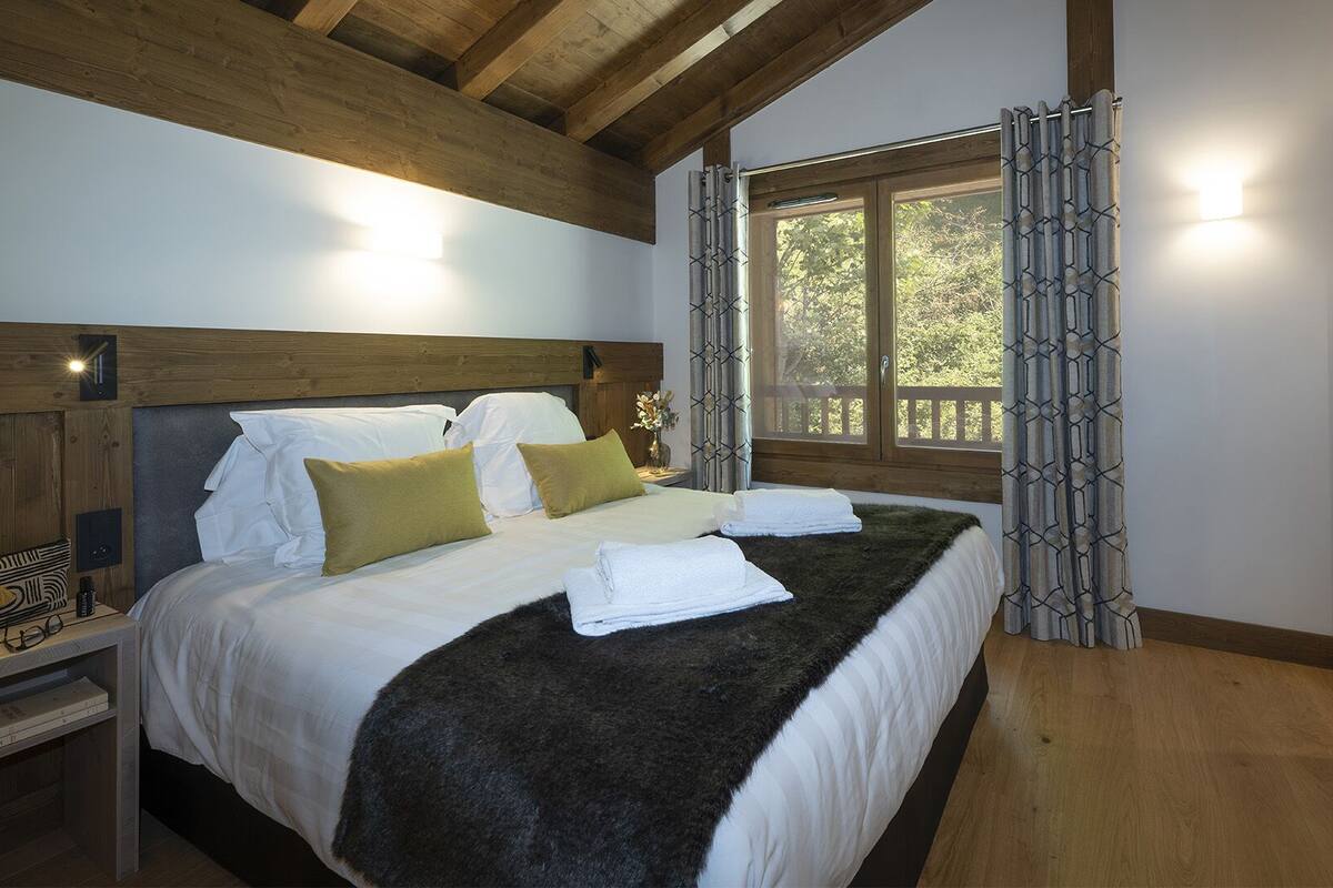 A bedroom with a wooden headboard, a large window with a view of trees, and a bed with white linens and a dark throw.