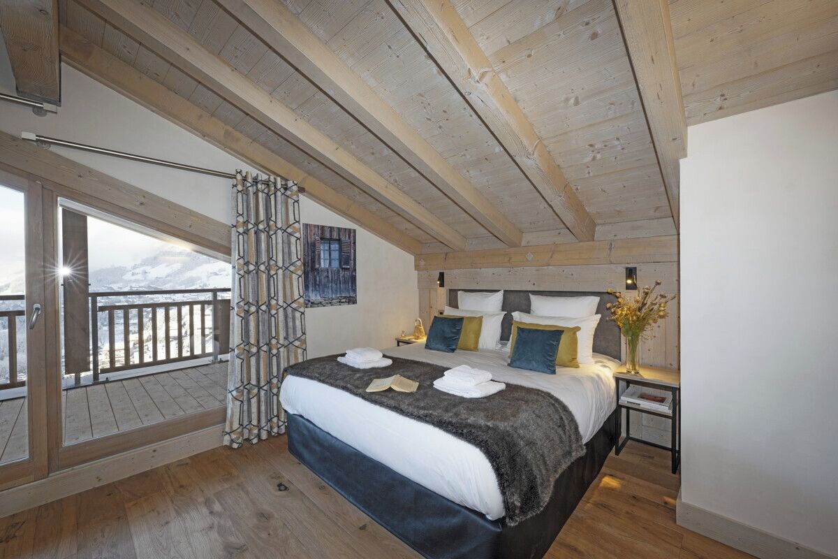 Quarto com cama grande, vigas de madeira, varanda com vista para montanhas e janela com cortinas.