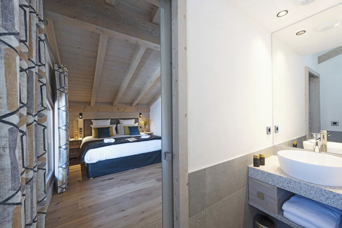 A bedroom with a large bed, a bedside table, and a nightstand. There is a bathroom with a sink, mirror, and towel rack.