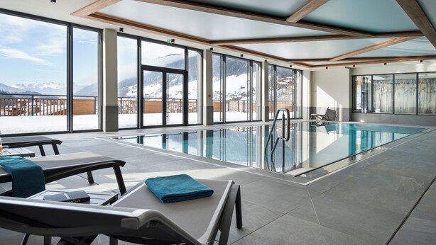 A modern indoor swimming pool with clear glass walls, wooden beams, and a view of snow-capped mountains. - MGM Hôtels & Résidences – Résidence Chalets de Joy (Le Grand-Bornand)