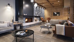 A modern living room with a fireplace, sofas, a coffee table, and a television mounted on the wall. - MGM Hôtels & Résidences – Résidence Chalets de Joy (Le Grand-Bornand)