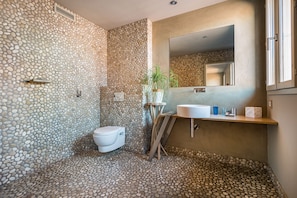 Un baño con piso y paredes de diseño con motivos de guijarros, un inodoro blanco, un lavamanos con espejo y una ventana.