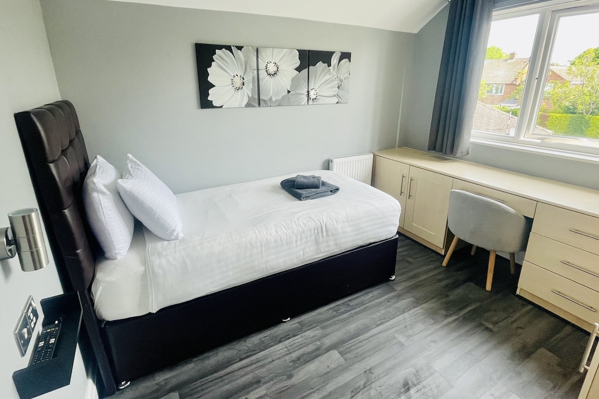 A bedroom with a bed, a desk, and a chair. There is a window with curtains, and a painting on the wall.