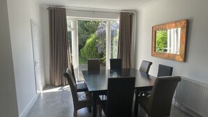 A dining area with a table and chairs, large windows with curtains, and a view of greenery outside. - Regency House (Loughborough)