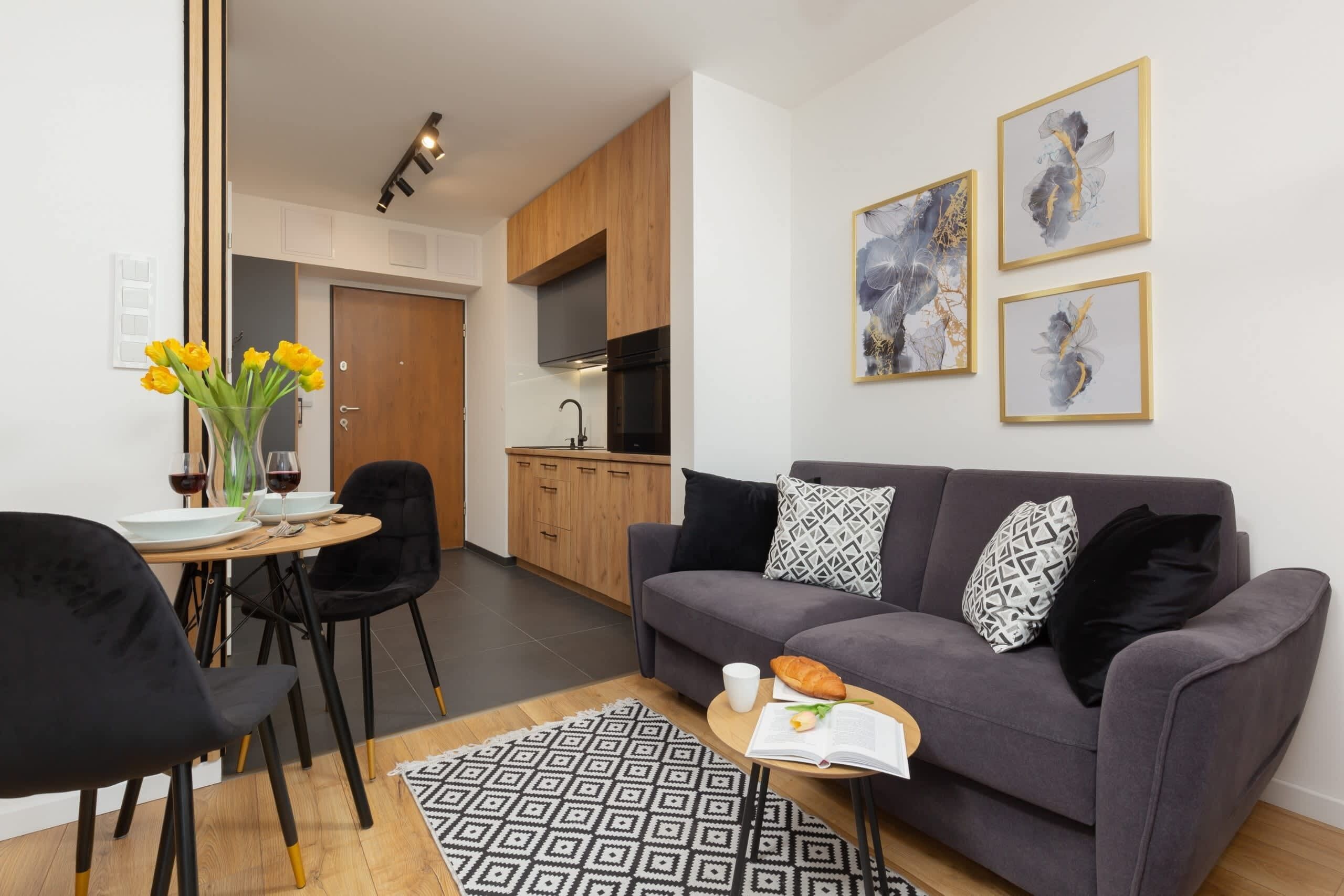 A modern living room with a grey sofa, black chairs, a dining table with tulips, and a kitchen in the background.