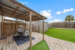Un patio cubierto con una mesa y sillas, rodeado por una cerca de madera y un césped cuidado.