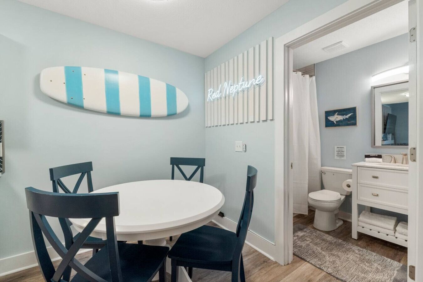 Una habitación con una mesa redonda y sillas, un adorno con forma de tabla de surf, un letrero de neón, una cortina de ducha y un baño con inodoro y lavamanos.