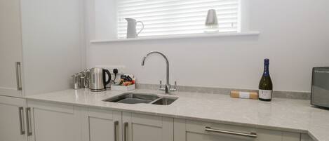 A modern kitchen with white cabinets, a double sink, a kettle, a wine bottle, and a small refrigerator.