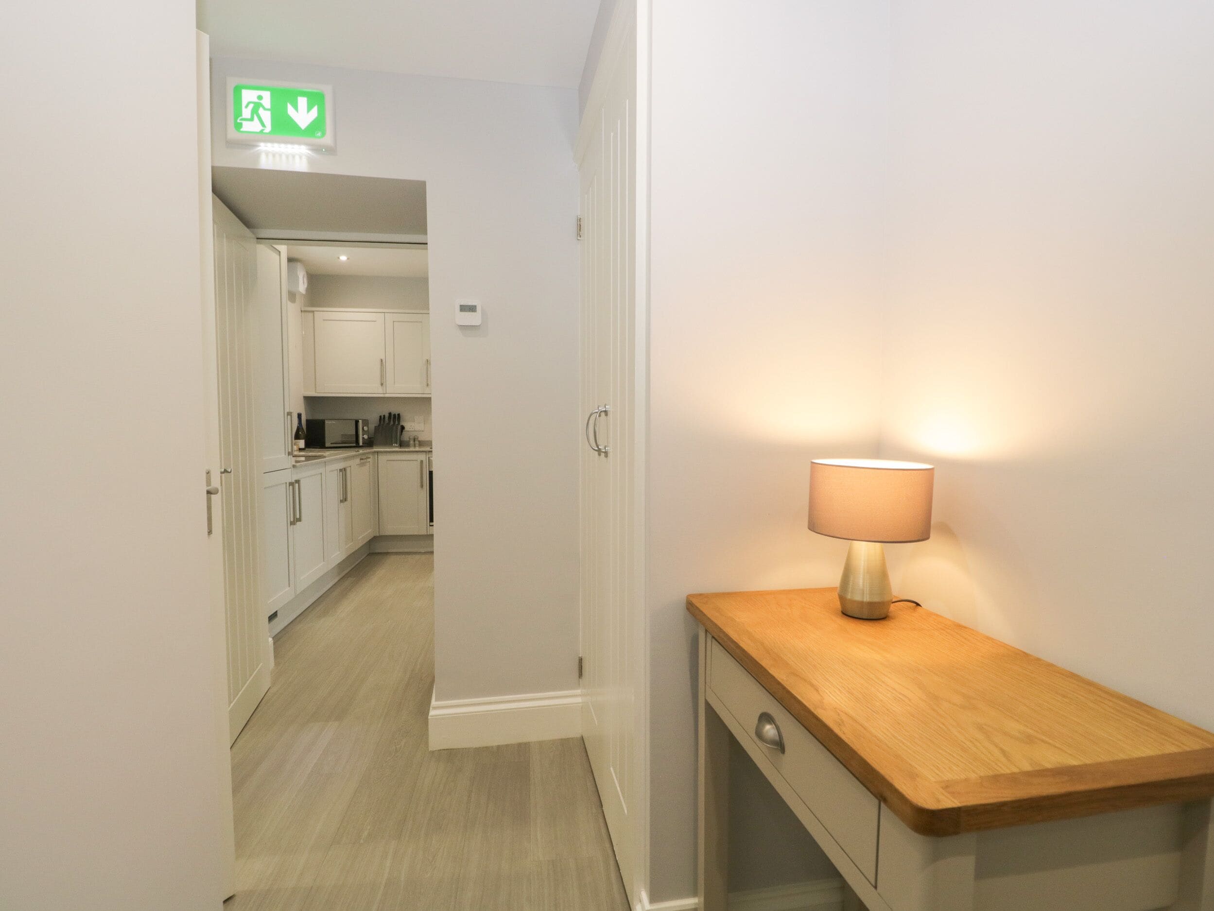 A hallway with a wooden nightstand and a lamp, leading to a kitchen area with white cabinets and a microwave.