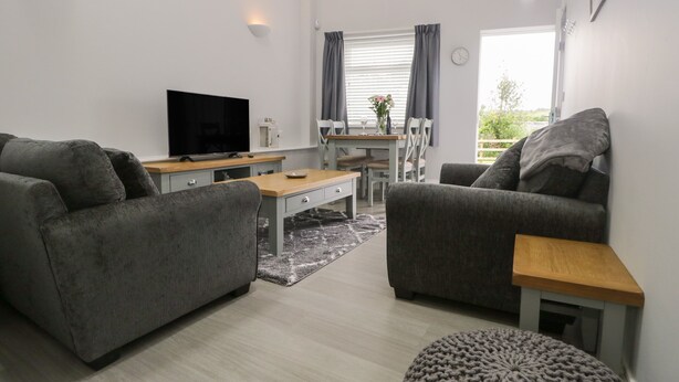 Un salon moderne avec deux canapés gris, une table basse en bois, un téléviseur à écran plat, un coin repas avec une table et des chaises blanches, et une fenêtre avec des rideaux.
