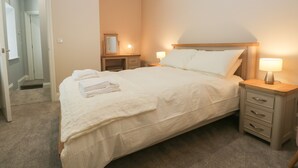 Un lit bien fait, avec du linge de lit blanc et une tête de lit en bois, deux tables de chevet avec des lampes, un miroir au-dessus de la commode et une porte donnant sur une autre pièce.