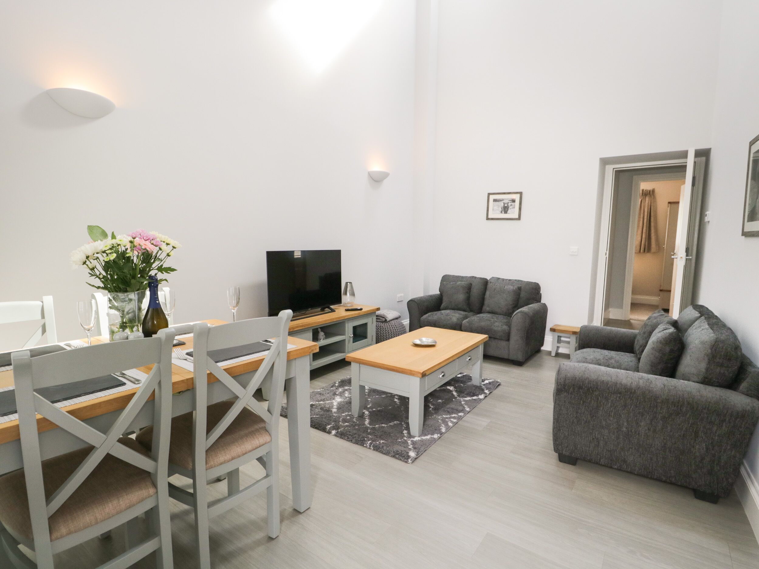 A modern living room with a dining area, grey sofas, a flat-screen TV, and a vase with flowers.