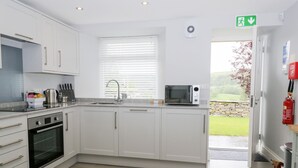 A modern kitchen with white cabinets, a built-in oven, a microwave, and a window with blinds. - Oxen Park Farm Cottage (Ulverston)