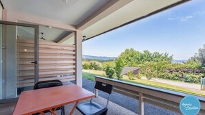 Un balcón con mesa y sillas, una puerta corrediza de vidrio y vistas a un jardín y colinas distantes.
