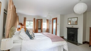 A bedroom with a bed, a bedside table, a lamp, a mirror, a fireplace, and a window with curtains.