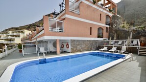 A swimming pool with lounge chairs and umbrellas, surrounded by a building with balconies and a stone wall. - Apartment with communal swimming pool in Playa de Mogan - Paraiso (lomo quiebre)