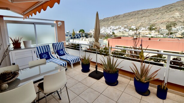 A terrace with a table and chairs, potted plants, and a view of a hillside town. - Apartment with communal swimming pool in Playa de Mogan - Paraiso (lomo quiebre)