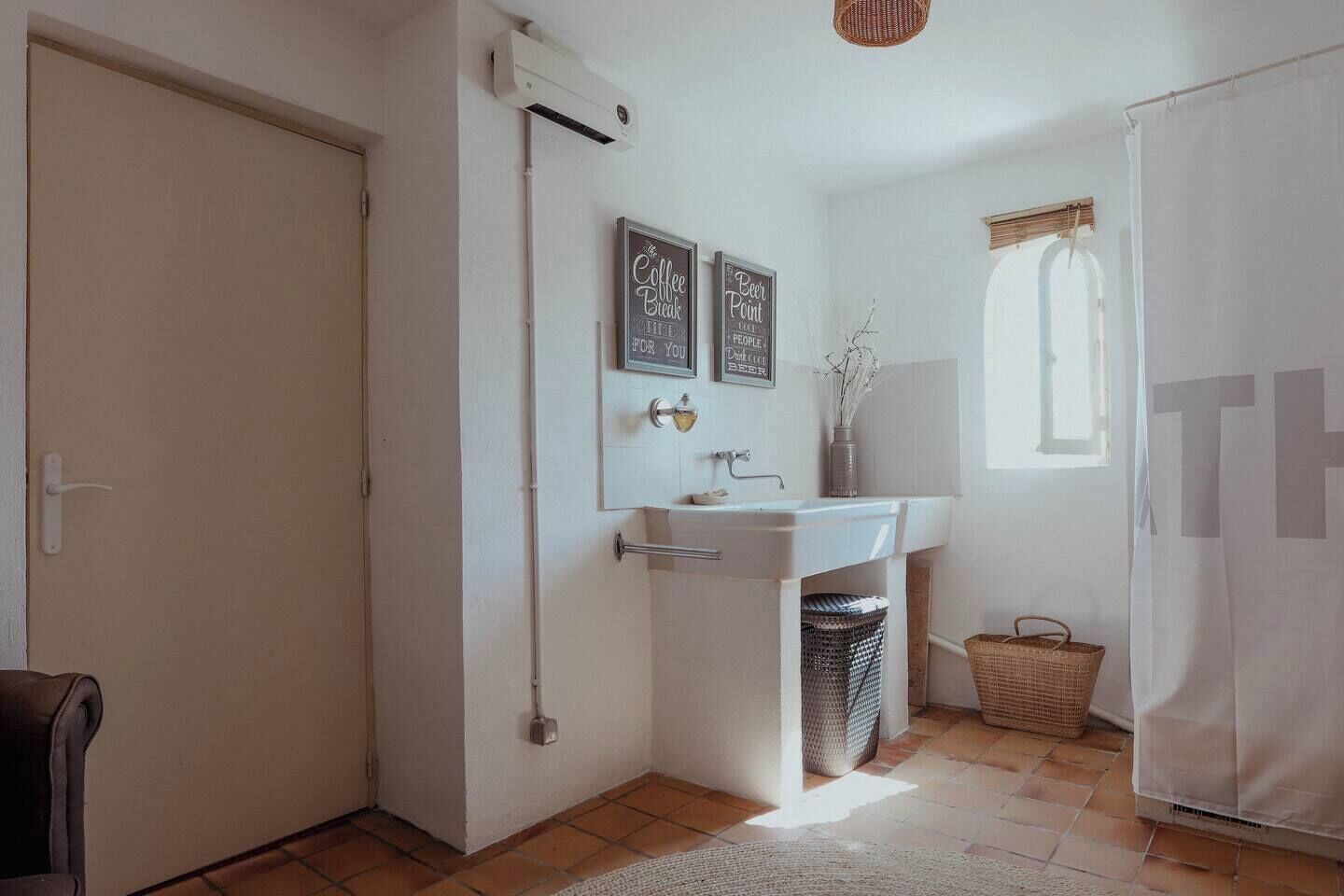 Un baño con un lavamanos blanco, un espejo y dos carteles enmarcados en la pared. Se ve una ventana con vistas al exterior, una puerta y una cesta en el suelo.