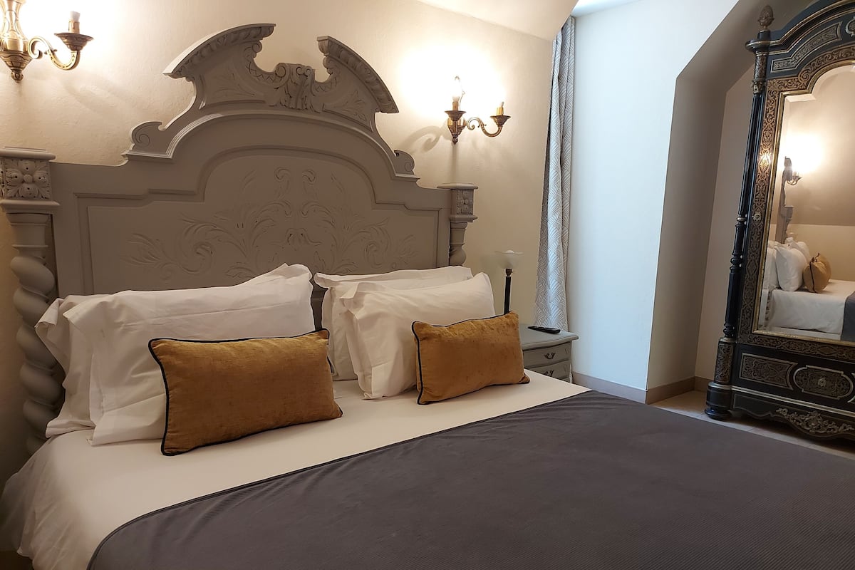 Cama bem arrumada com travesseiros brancos e castanhos, edredom cinza escuro e cabeceira decorativa com arandelas.