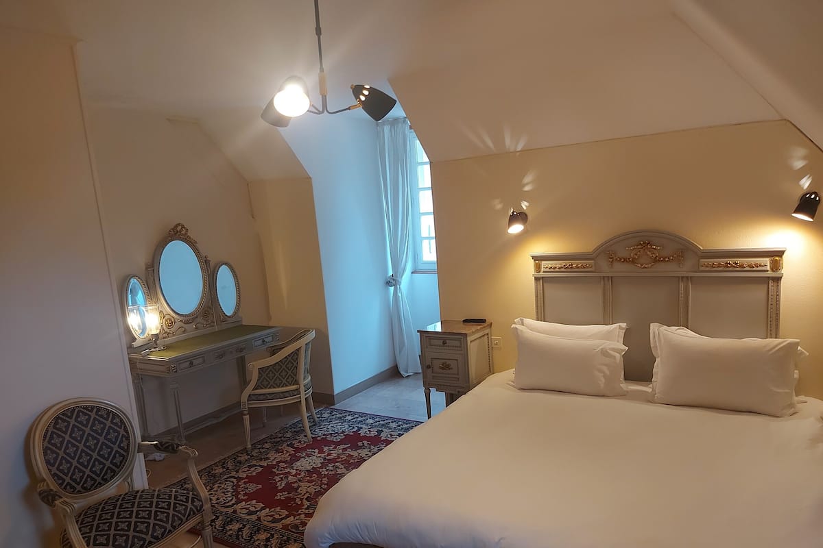 Quarto com cama grande, penteadeira com espelhos, uma cadeira e mesa de cabeceira.