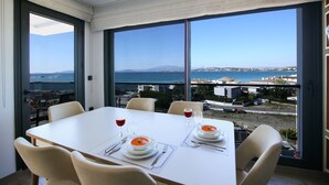 A dining area with a table set for two, large windows offering a view of the sea and buildings. - Krem Marin Suites (Cesme)