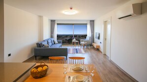 A modern living room with a sofa, dining table set, and a view of the city through large windows. - Krem Marin Suites (Cesme)
