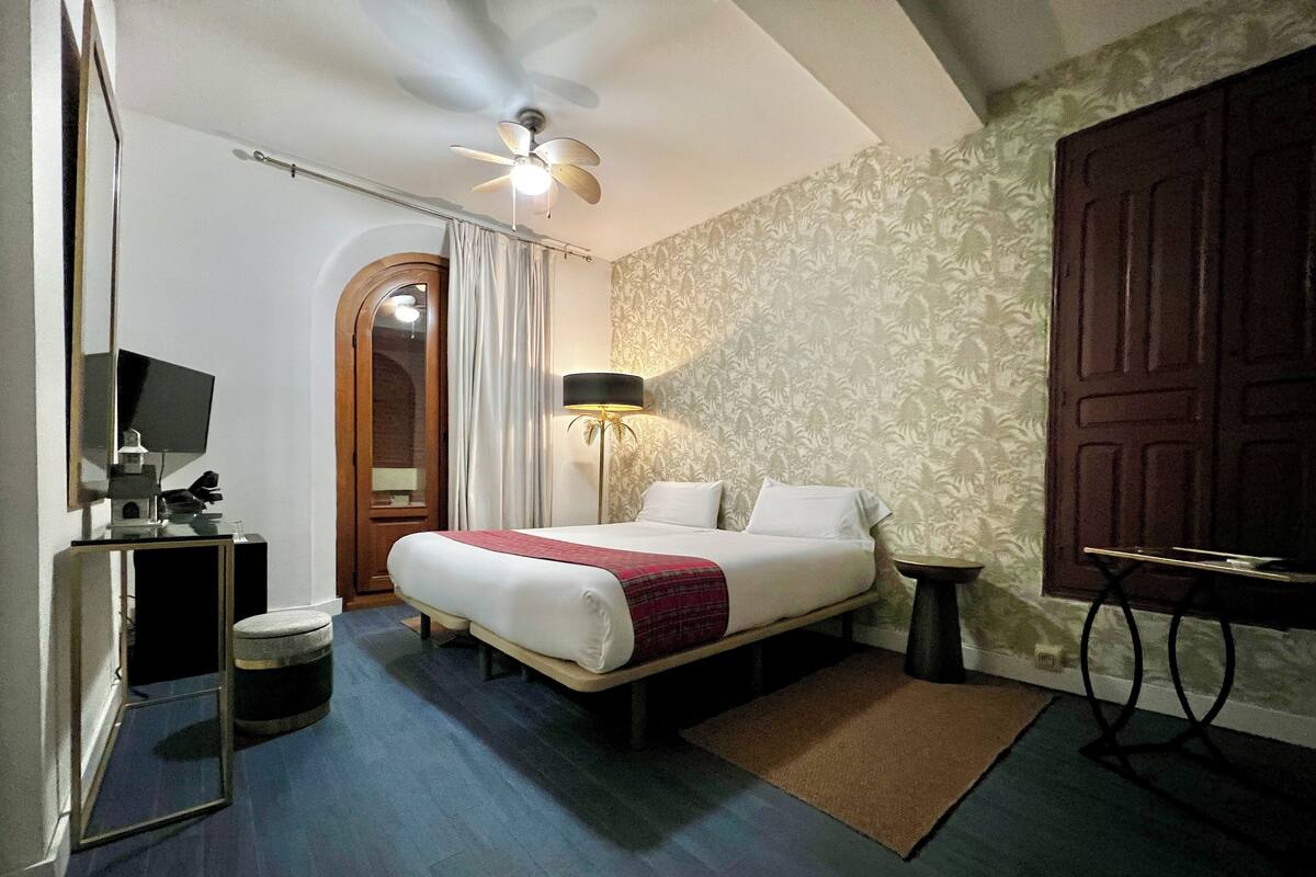 A hotel room with a bed, a desk with a computer, a ceiling fan, a lamp, and a door.