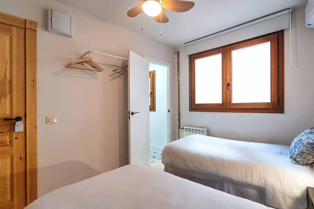 A bedroom with two beds, a wooden wardrobe, a ceiling fan, a window with wooden frames, and a radiator.