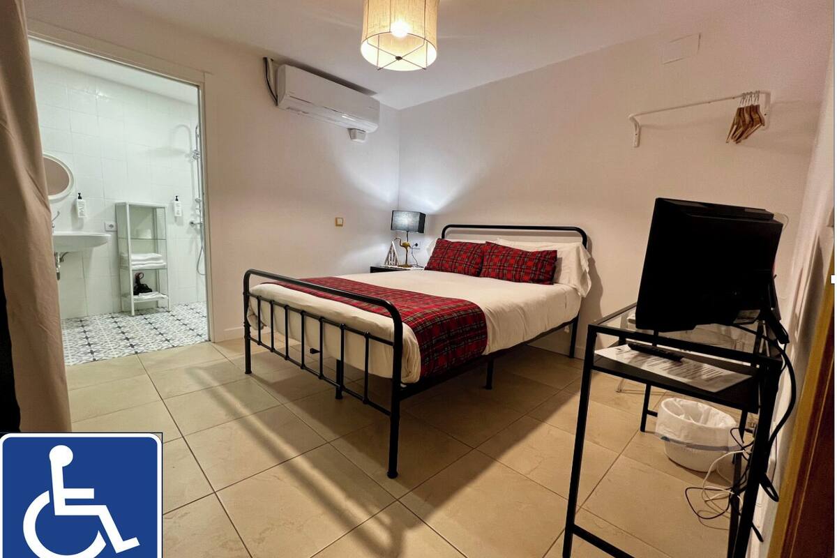 A bedroom with a bed, a TV on a stand, a desk, and a bathroom visible through an open door.