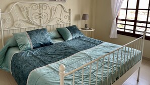 Un llit amb capçalera metàl·lica, una tauleta de nit amb una làmpada i una finestra amb cortines.