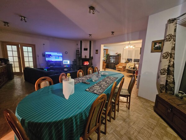 A dining area with a table set for six, surrounded by wooden chairs. There is a television in the background, and a fireplace is visible in the adjacent room.