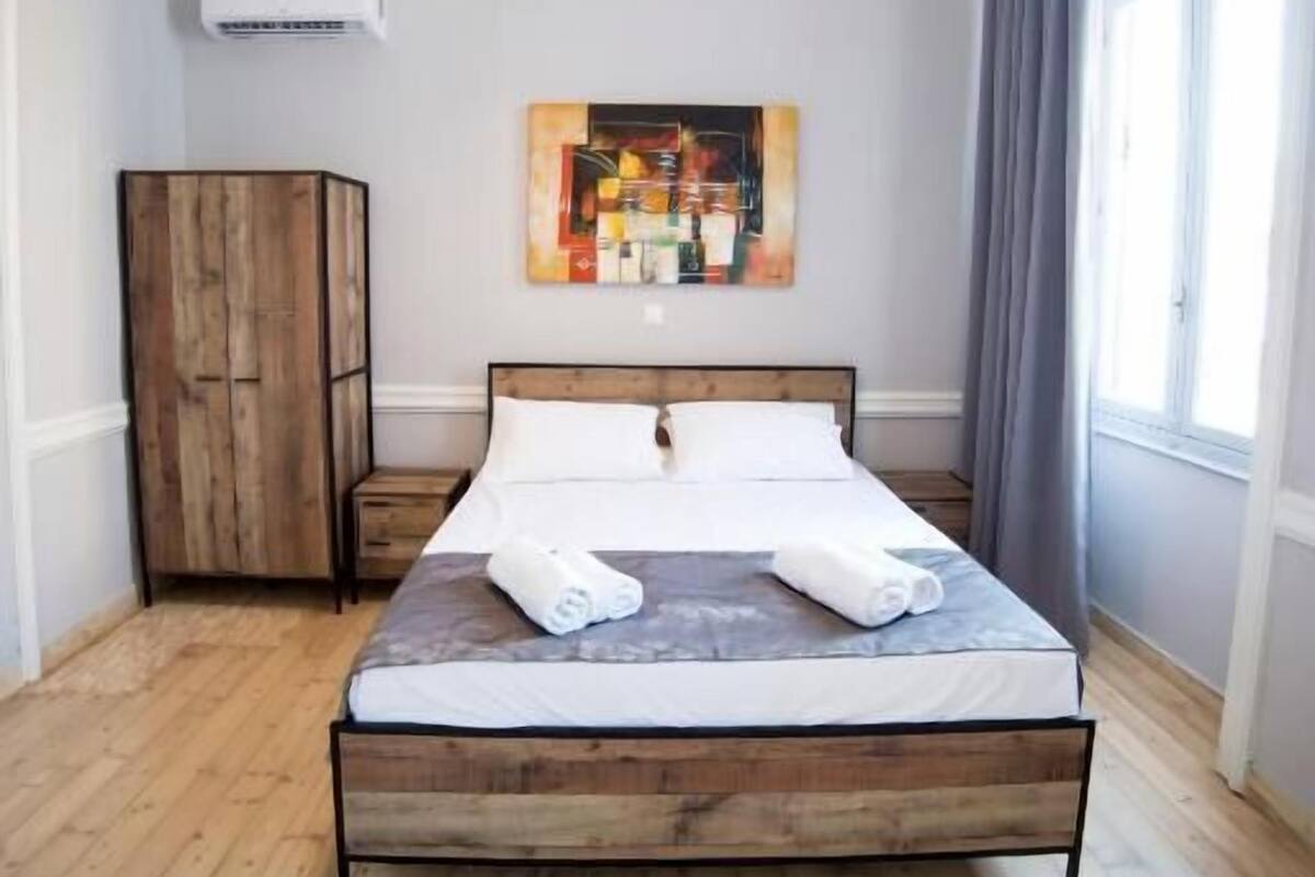 Ein Schlafzimmer mit einem hölzernen Bett, einem Kleiderschrank, einem Bild an der Wand und einem Fenster mit Vorhängen.