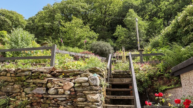 Uma escadaria de pedra leva a uma cerca de madeira, com um jardim e árvores ao fundo.