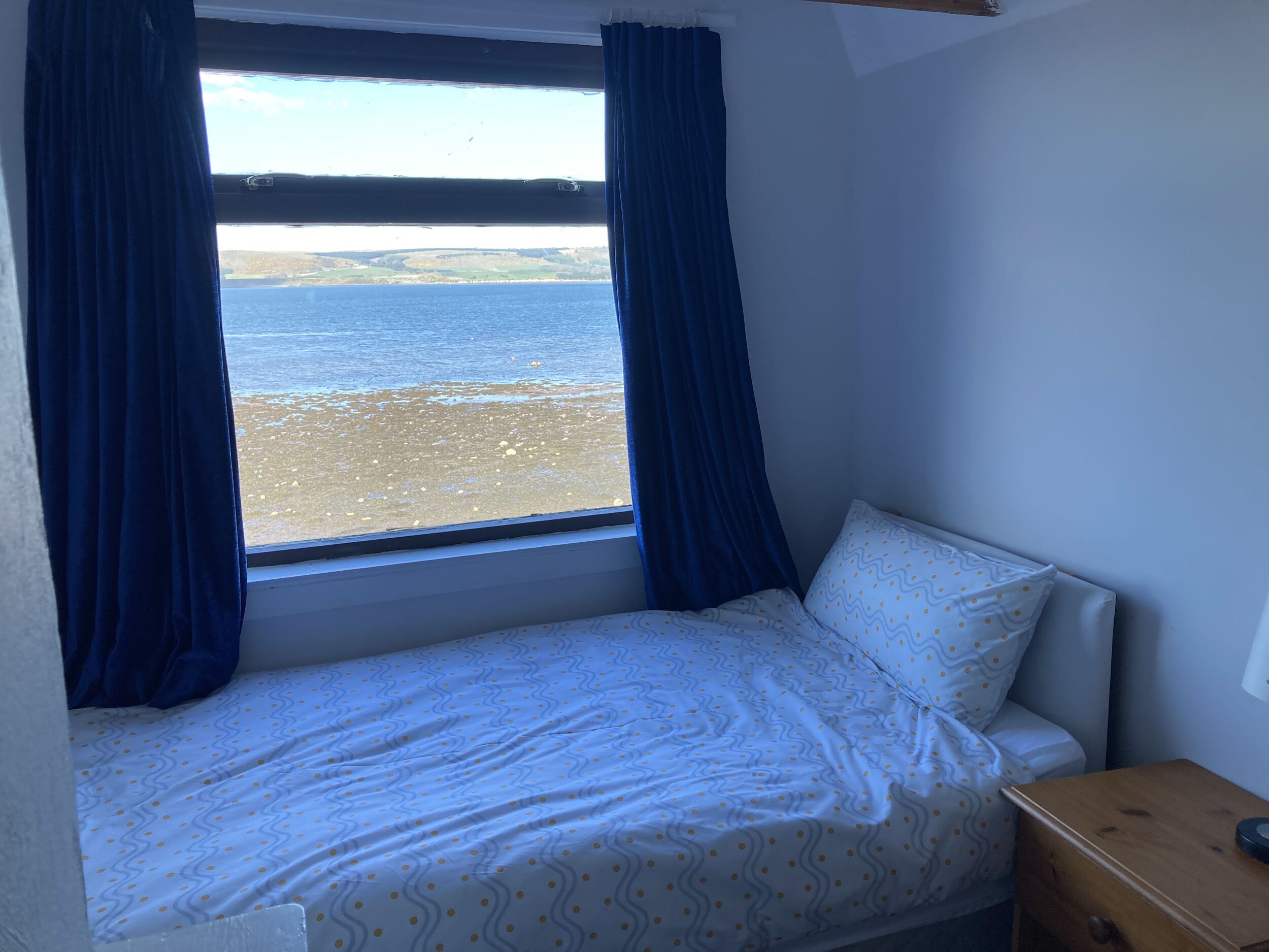 A small bedroom with a single bed, a wooden nightstand, and a window with curtains.