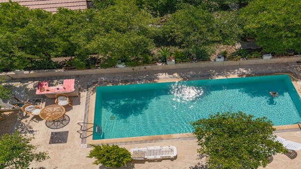 An aerial view of a rectangular swimming pool with a tiled deck, surrounded by greenery and outdoor furniture.