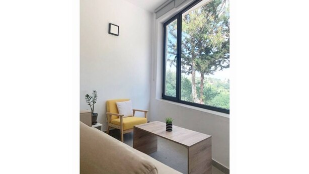 Un salon moderne avec un canapé beige, un fauteuil jaune, une petite table basse en bois et une grande fenêtre donnant sur des arbres.