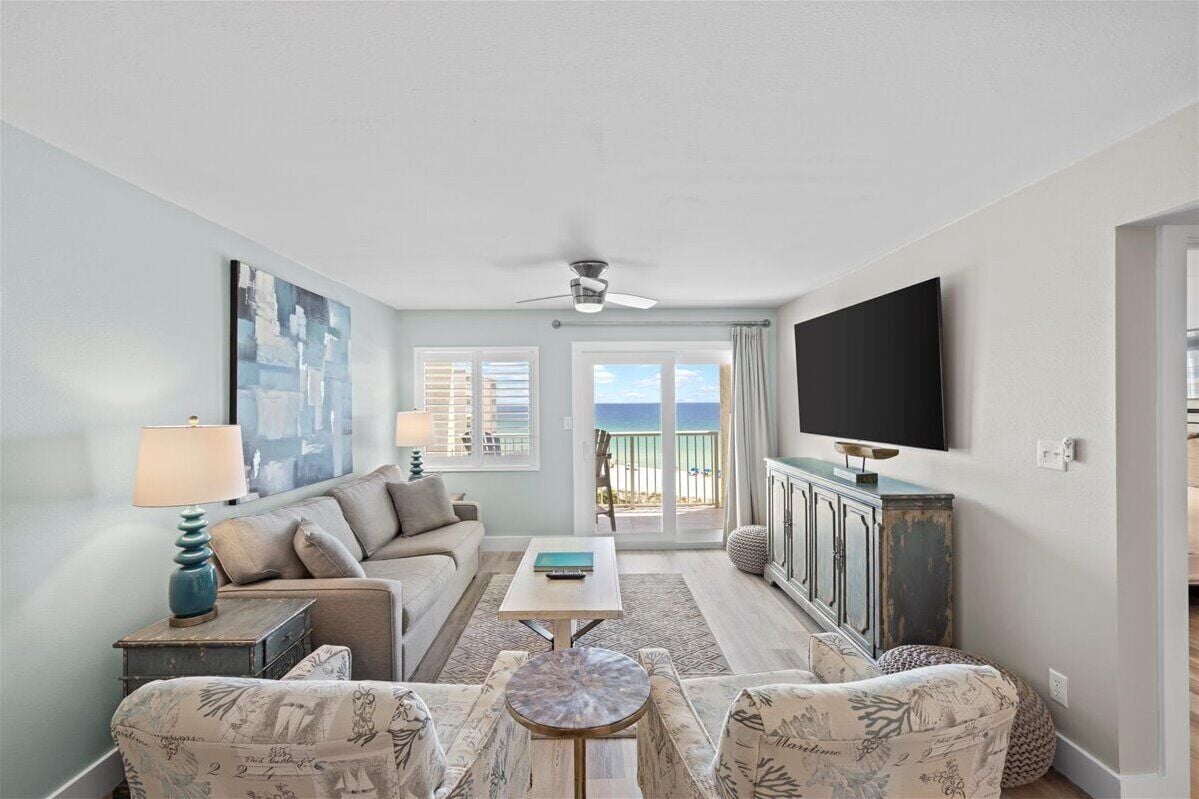 A living room with a sofa, armchairs, a coffee table, a TV, and a view of the ocean through large windows.