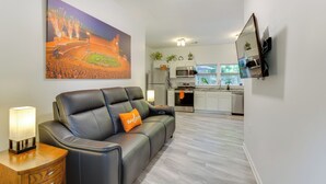 A modern living room with a leather sofa, a painting of fireworks, a TV, and a kitchen in the background. - Clemson Game Day Cottage w/ Fire Pit (Central)