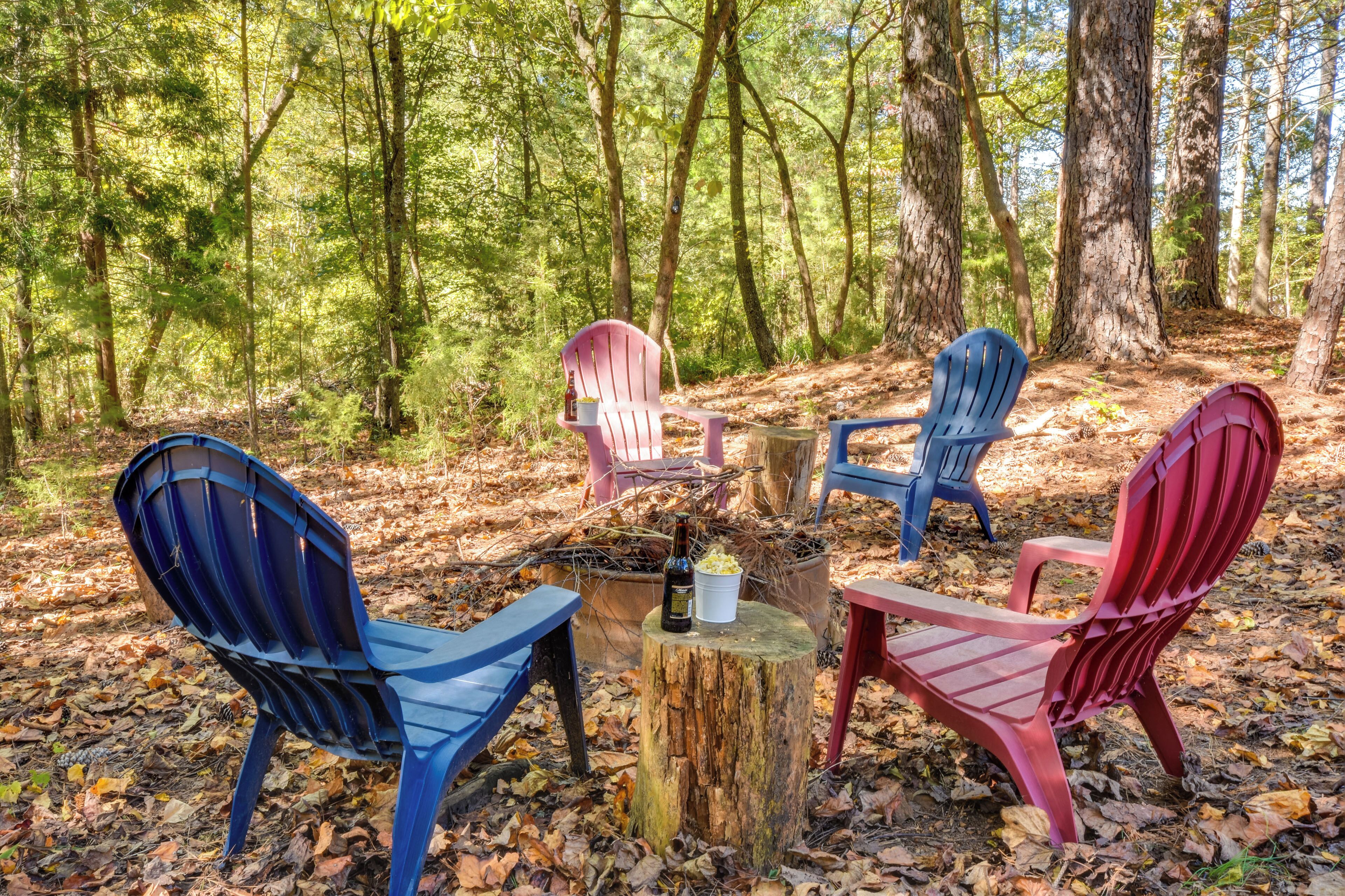 A group of colorful Adirondack chairs arranged around a wooden stump in a forest setting.