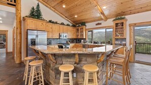 A kitchen with wooden cabinets, a central island with stools, a stone backsplash, and a view of the outdoors. - The Golden Oak Retreat (Big Bear Lake)