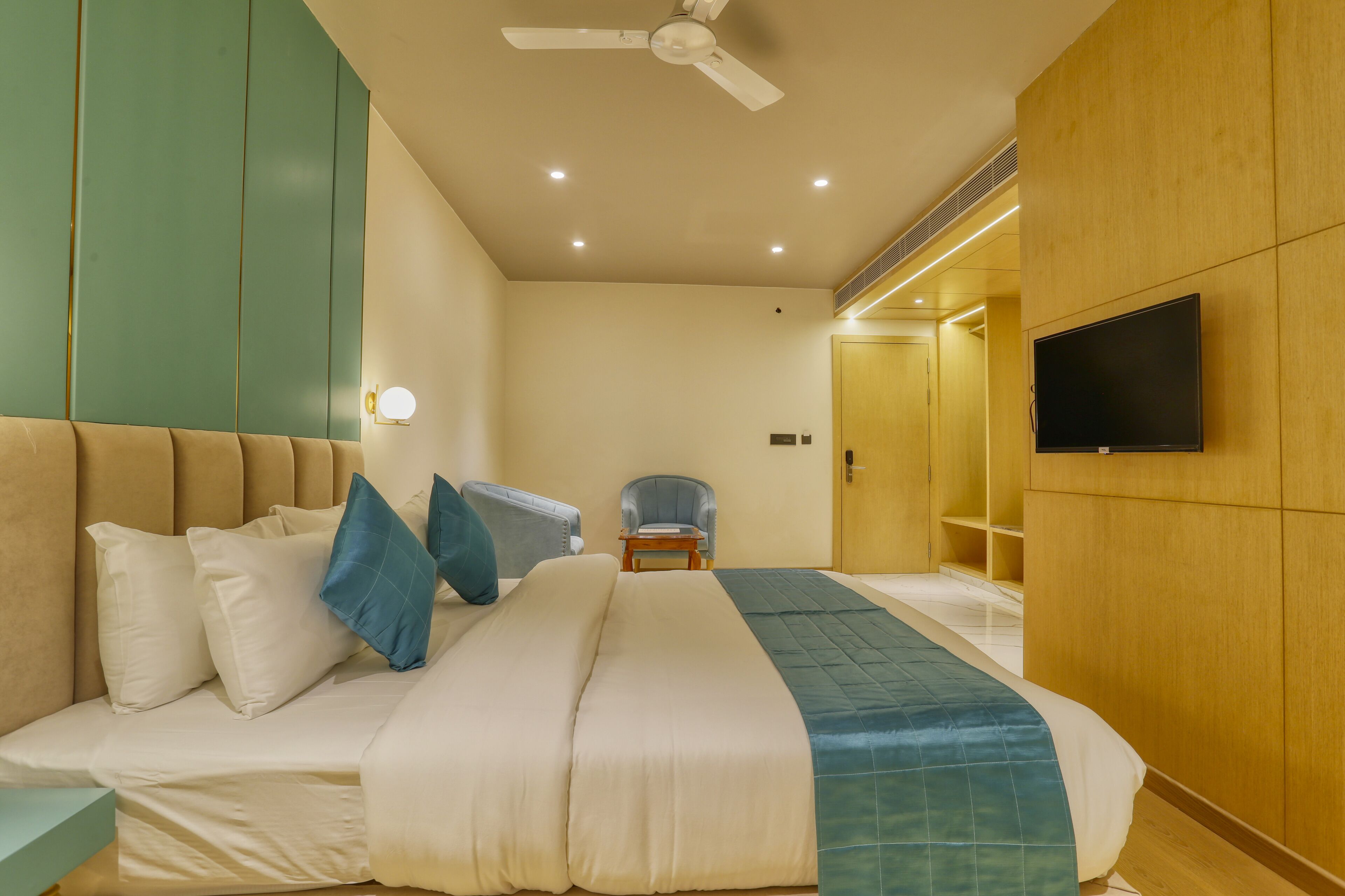 A hotel room with a large bed, a ceiling fan, a TV mounted on the wall, and a small seating area with a chair and table.