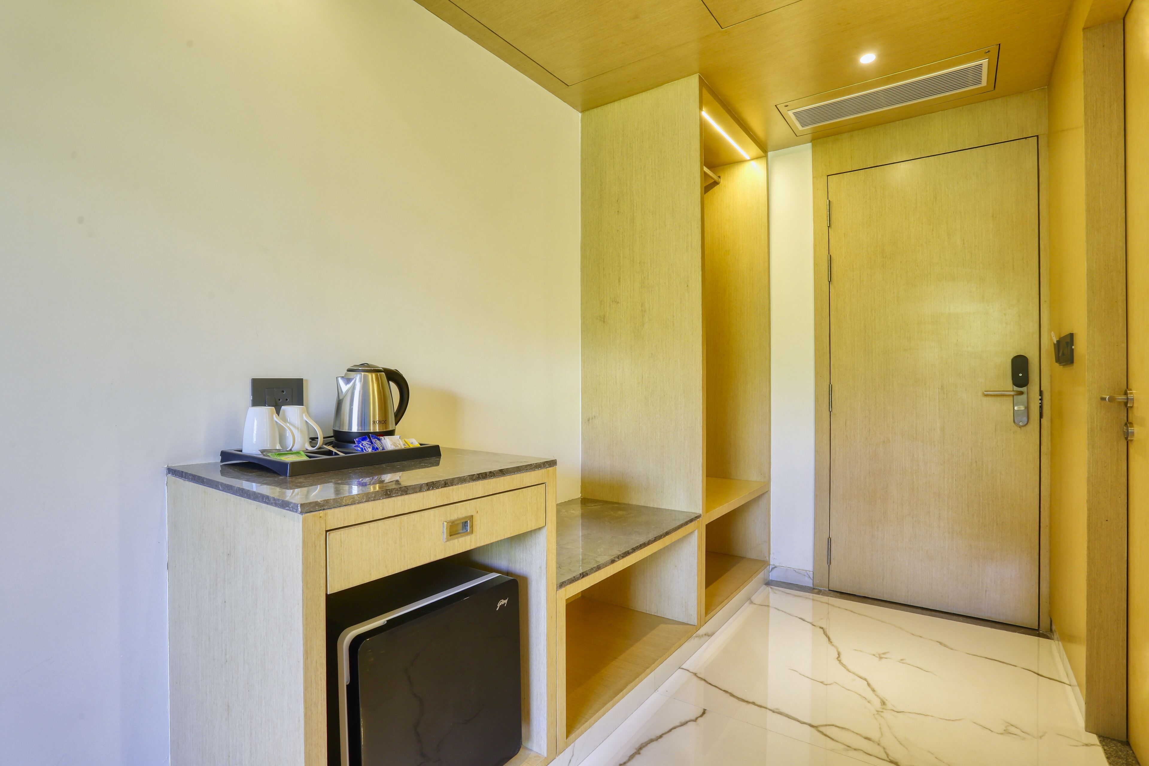 A compact hotel room with a wooden desk, a small refrigerator, a door with a handle, and a marble floor.