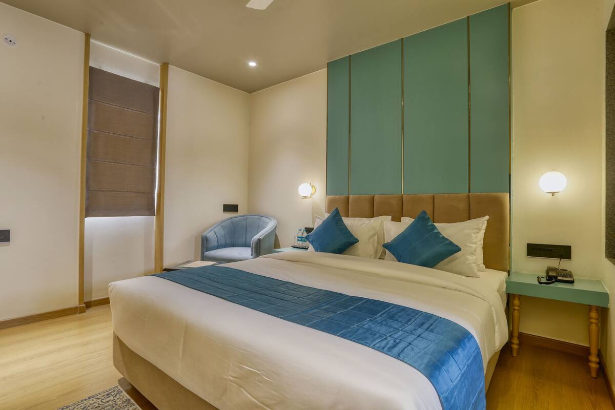 Ein ordentlich eingerichtetes Hotelzimmer mit einem großen Bett, einem Sessel, einem Nachttisch mit Lampe und einem an der Wand befestigten Telefon.