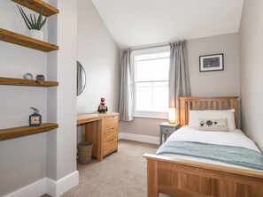 A bedroom with a wooden bed, a desk with drawers, a small shelf with decorative items, a window with curtains, and a framed picture on the wall.