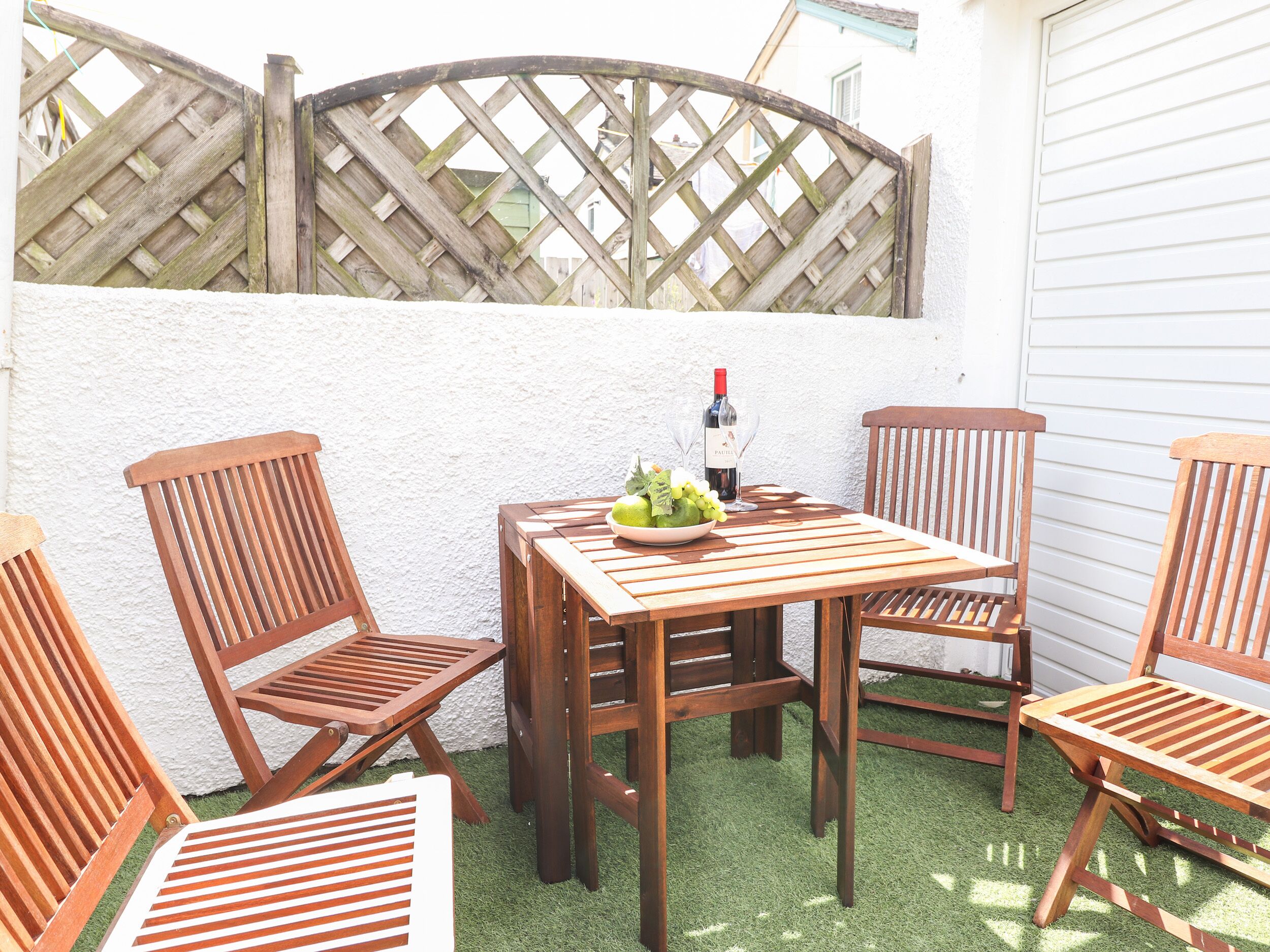 A wooden table with chairs, a bowl of fruit, and a bottle of wine on a patio with artificial grass.