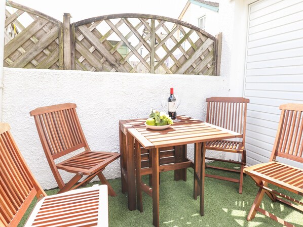 A wooden table with chairs, a bowl of fruit, and a bottle of wine on a patio with artificial grass.