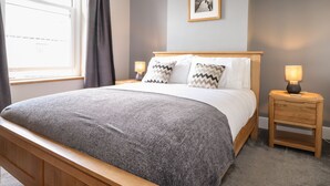 A neatly made bed with a grey blanket, two bedside tables with lamps, a framed picture on the wall, and a window with curtains. - Dovestones (Keswick)