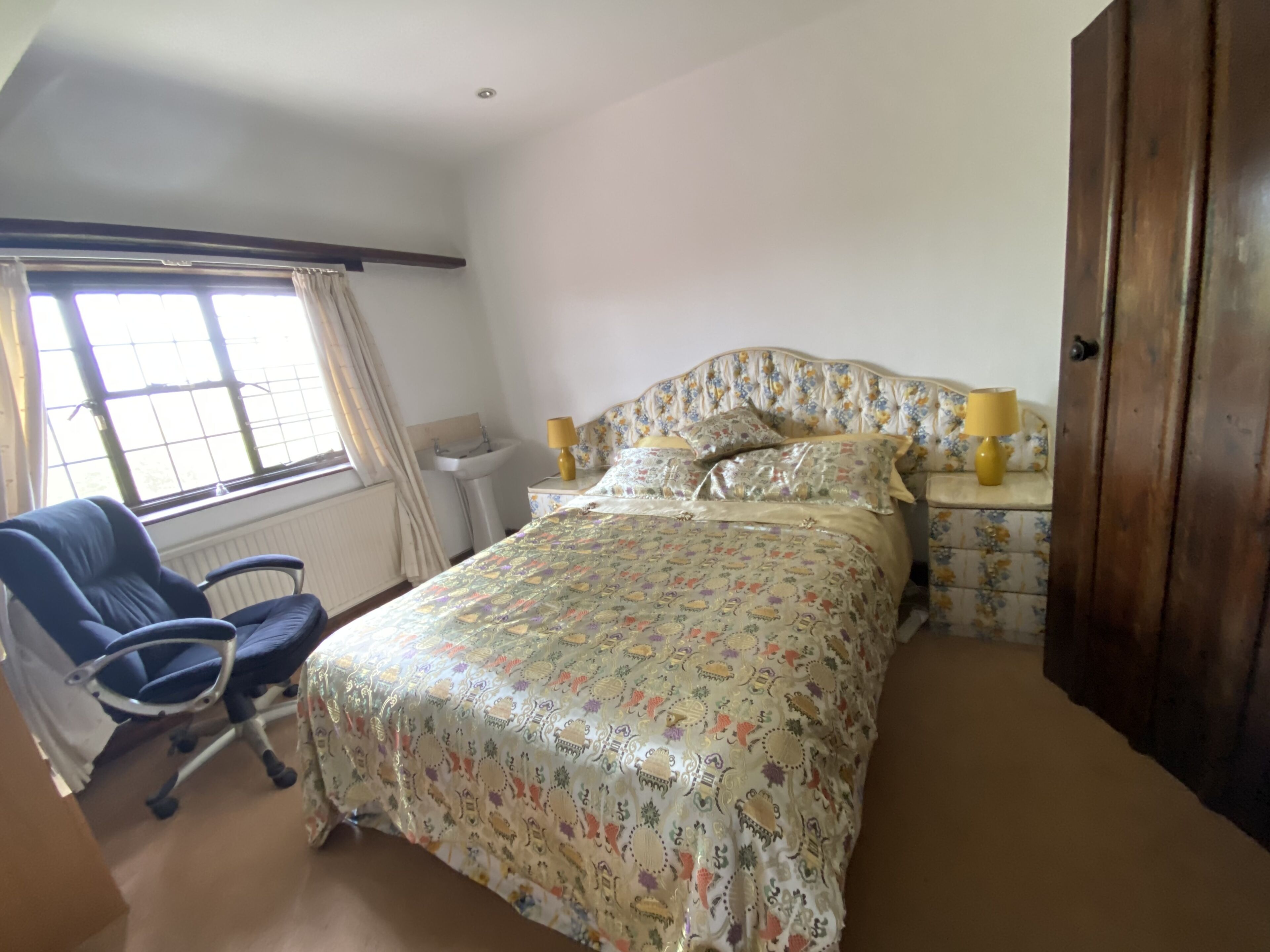 Un dormitorio con cama, una silla, una mesita de noche, lámpara, ventana con cortinas y un radiador.